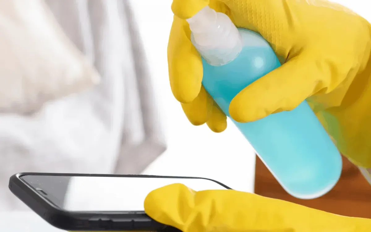 Sanitizing smartphone screen with disinfectant spray and yellow gloves.