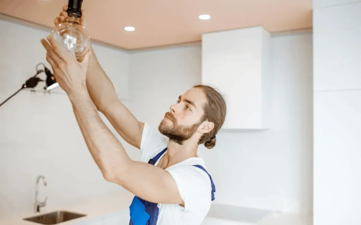 Male cleaner in uniform inspecting and cleaning a ceiling light bulb in a modern kitchen.