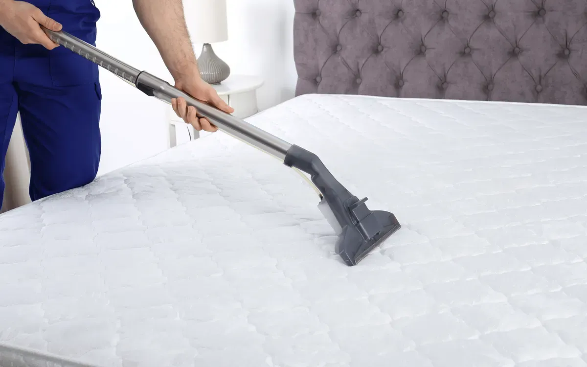 A cleaner in blue uniform using a steam vacuum cleaner on a white mattress in a bedroom.