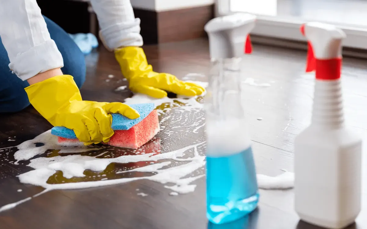 Person wearing yellow gloves scrubbing wooden floor with sponge and cleaning solution during professional home cleaning.