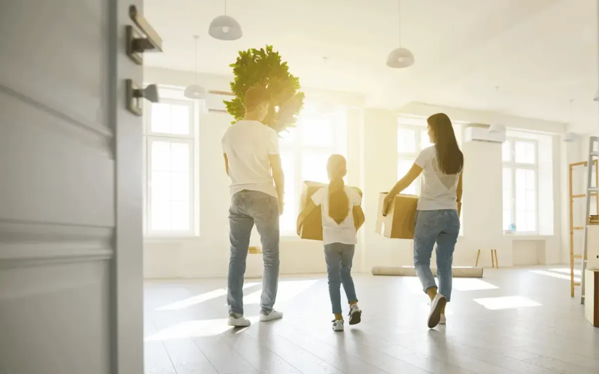 Family carrying boxes and a plant while moving into a bright new home.