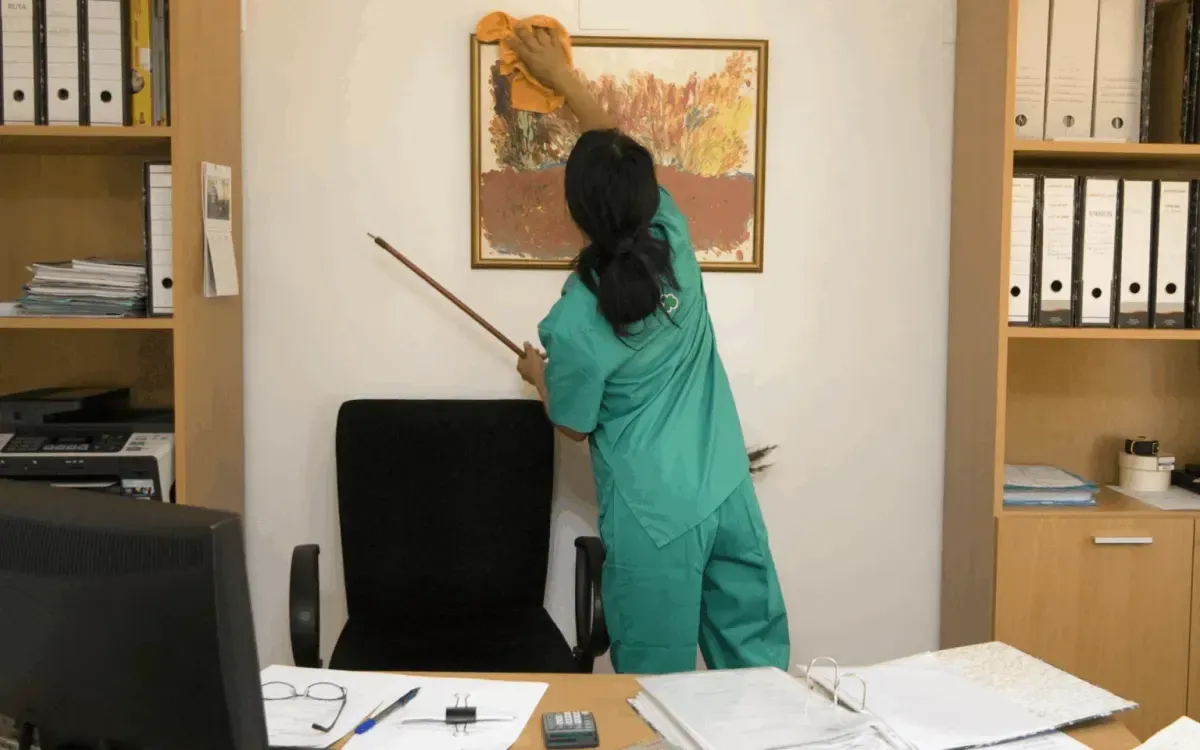 Professional cleaner dusting and wiping a picture frame in an office workspace.